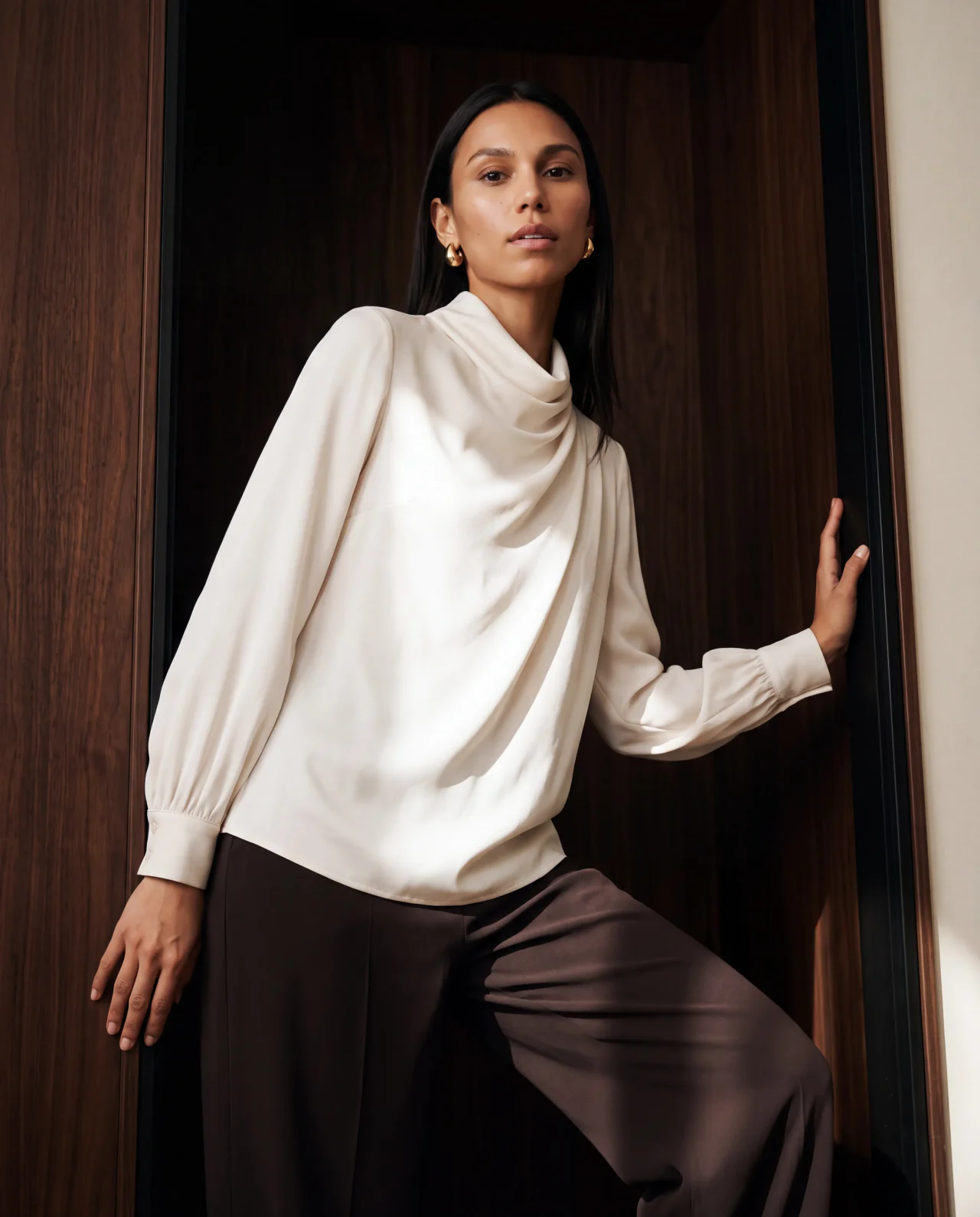 Sandra posing in the ivory draped blouse and espresso trousers against a walnut backdrop