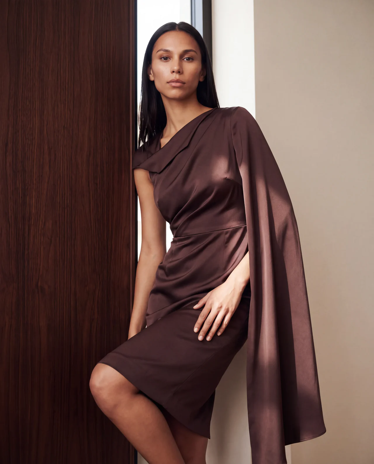 Sandra leaning against dark walnut in a draped cocoa dress